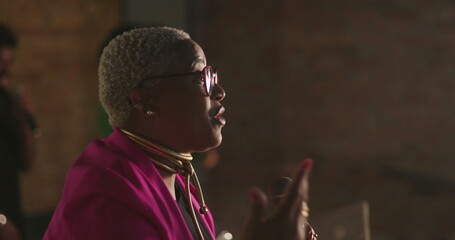 Evangelical preacher passionately addressing congregation, close-up profile, silver-haired Afro-descendant woman with glasses and pink jacket, powerful sermon delivery