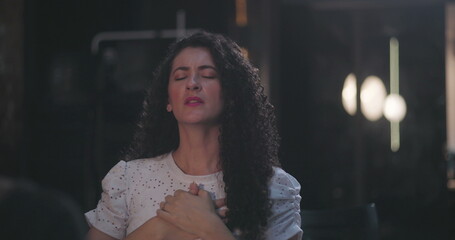 Curly-haired woman expressing gratitude, eyes closed, hands pressed against chest during peaceful...