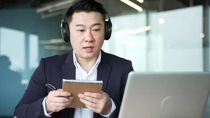 Asian businessman in headphones watching video call conference using a laptop takes notes in notebook in business office. Manager in a formal suit communicates remotely at an online meeting. Close up - Powered by Adobe