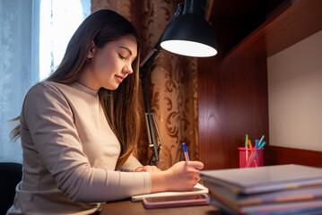 Teenage girl writing homework in notebook at desk in cozy room. A teenage girl doing homework at...