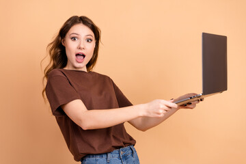 Surprised young woman holding laptop wearing casual brown shirt in front of beige background