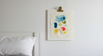 Bright watercolor painting with floral elements hung on a white wall near a bed