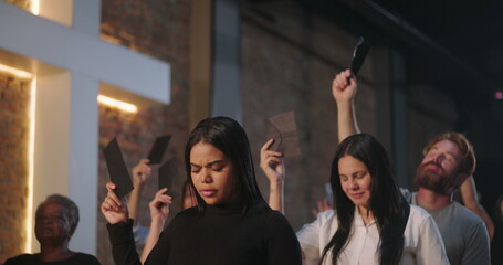 Diverse congregation raising offering envelopes during worship in church, illuminated cross on brick wall, spiritual unity, prayerful gathering, symbolic act of faith and devotion