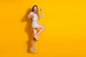 Full-length portrait of a stylish woman jumping with excitement against a vibrant yellow background, wearing a polka-dot outfit