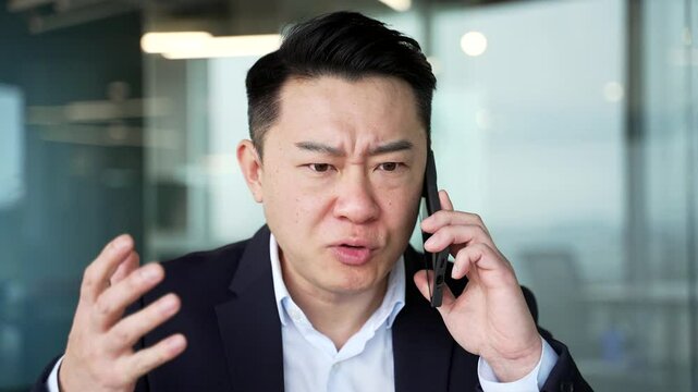 Angry asian businessman arguing talking on mobile phone at workplace in office. Excited nervous manager solving stressful business situation or conflict resolution. Boss scolds subordinate. Close up