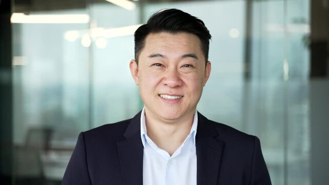 Portrait of smiling asian businessman in formal suit standing at workplace in modern business office. Head shot confident happy successful entrepreneur manager looking at camera in workspace. Close up