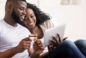 Cheerful Black Spouses Shopping Online Using Digital Tablet At Home, Entering Credit Card Security...