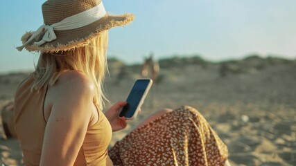 Blonde woman relaxing in flowy summer dress, wearing sunhat, scrolling smartphone while reclining on golden sandy beach during serene vacation.Summer concept.Travel, blogging, weekend, relax - Powered by Adobe