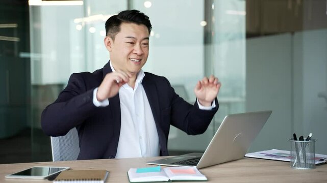 Happy asian businessman finished work on laptop sitting at desk at workplace in business office. Smiling satisfied man in formal suit puts his hands behind head, stretches himself in chair. Work done