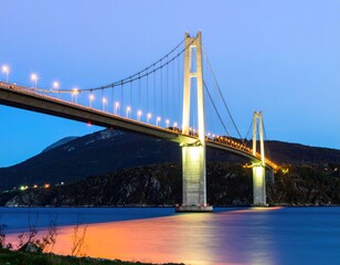Fototapeta premium A majestic cable-stayed bridge spans a tranquil waterway at twilight. Illuminated supports and roadway, with a softly lit, distant mountain range