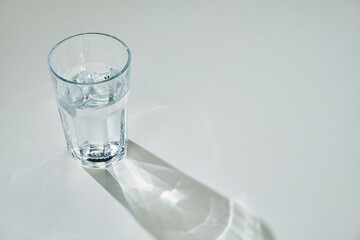 Clear glass of water casting light reflections and a shadow on a white surface. Minimalist still life with bright lighting and clean composition, emphasizing purity and hydration