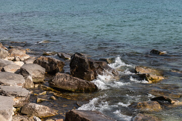 A natural lakeside scene with clear water and gentle waves breaking over large, weathered rocks along the shoreline.