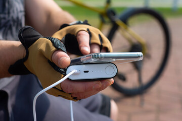 A male cyclist wearing sports gloves charges his smartphone with a portable power bank. The concept of modern technology and active lifestyle in nature, gadgets and energy on the go.