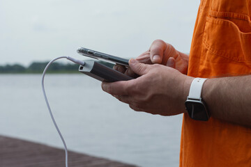 Man charging smartphone with power bank by lake using USB cable in summer outdoor. Concept modern lifestyle, mobile technology, travel, and connectivity on the go.