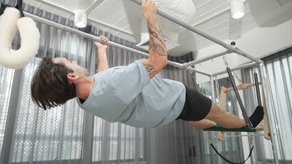 Man gripping bar on pilates trapeze table for core strength and upper body workout in a gym. Trainer hanging inverted back bend on the cadillac for improving flexibility in back and hip. Habituate.