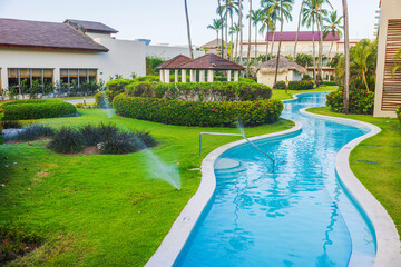 Curved swimming pool with clear water and sprinklers on green lawn surrounded by palm trees and hotel.  Dominican Republic. Punta Cana. 