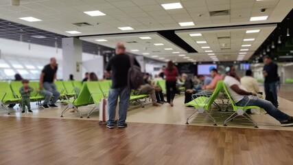 Long exposure time-lapse of people moving with motion blur in a modern airport terminal waiting area, surrounded by passengers sitting on green chairs under bright artificial lighting.