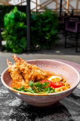 A bowl of ramen with tempura shrimp, boiled egg, cherry tomatoes, cilantro, and noodles in rich broth, set on a marble table outdoors with lush green plants in the background.