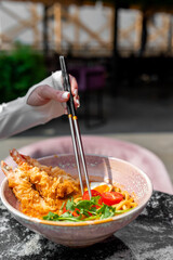 A close-up of a bowl of ramen with tempura shrimp, cherry tomatoes, and cilantro. A hand holding chopsticks is picking up noodles. The background is blurred, suggesting an outdoor dining setting