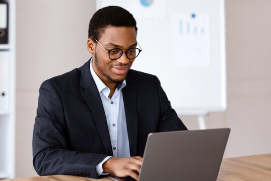 Fototapeta Smiling young black entrepreneur in formal suit working with laptop, wearing glasses, office interior, copy space. Successful millennial african american businessman working on business project