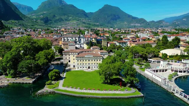 Aerial view of Riva del Garda with Torre Apponale, MAG museum, bike paths, boats and river ferries connecting towns, surrounded by mountains and clear Lake Garda water, full of tourists