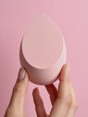 A hand grips a pink makeup sponge, set against a soft pink background, highlighting the simplicity and color harmony.