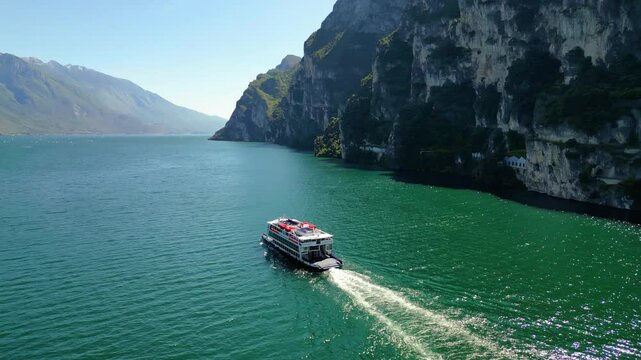 Aerial view of Riva del Garda with Torre Apponale, MAG museum, bike paths, boats and river ferries connecting towns, surrounded by mountains and clear Lake Garda water, full of tourists