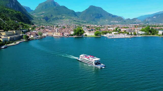 Aerial view of Riva del Garda with Torre Apponale, MAG museum, bike paths, boats and river ferries connecting towns, surrounded by mountains and clear Lake Garda water, full of tourists