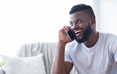Talkative african american man talking on phone with friends, sitting on sofa at home, free space