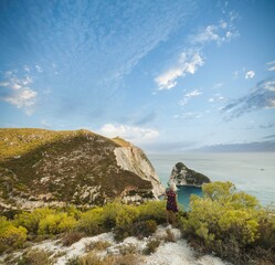 A traveler stands on a scenic cliff in Zakynthos, gazing at the azure waters and dramatic landscape. The vibrant summer sky enhances the serene atmosphere and natural beauty
