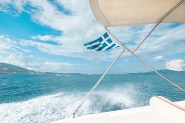 Tourists enjoy a serene boat ride in Zakynthos, with the vibrant Greek flag waving against a backdrop of shimmering blue seas and fluffy white clouds in summer