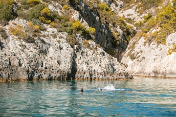 In the warm sunlight, two swimmers enjoy the crystalline waters of Zakynthos, surrounded by stunning rocky cliffs and lush greenery. A perfect summer escape awaits