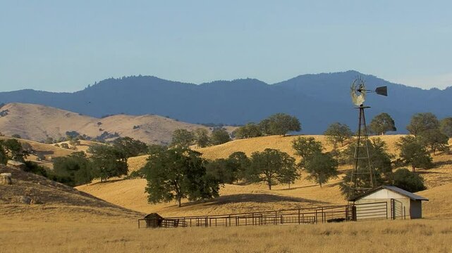 A corral, outbuilding and a water windmill set in a golden grass field are set before oak-studded hills and a background of pine-covered mountains in the foothills of California's Sierra Nevada.