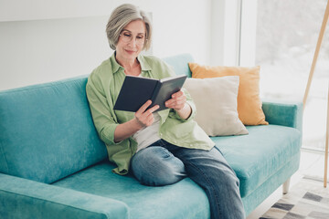 Obraz premium Mature Woman Relaxing on a Comfortable Sofa Reading a Book at Home in a Well-Lit Living Room