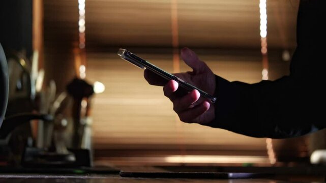 A close-up shot captures the modern and connected mood as a hand holds a smartphone, silhouetted against warm light filtering through blinds, suggesting communication or digital interaction
