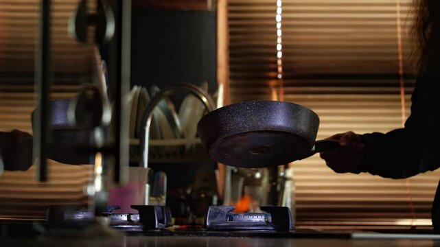 A dynamic mid-shot captures a focused mood as a hand holds a frying pan over a gas stove burner, poised for cooking in a warm, dimly lit kitchen, suggesting culinary action