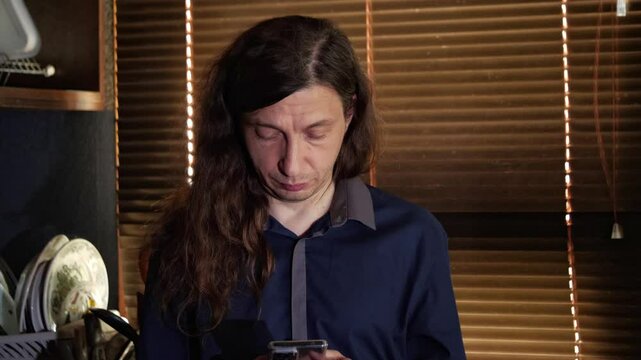 A mid-shot captures a curious mood as a man with long hair looks at his smartphone, with a distinct blue light reflecting on his face, suggesting a video call or content viewing in a dimly lit room