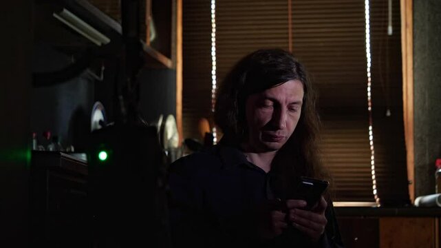 A mid-shot captures a curious mood as a man with long hair looks at his smartphone, with a distinct blue light reflecting on his face, suggesting a video call or content viewing in a dimly lit room