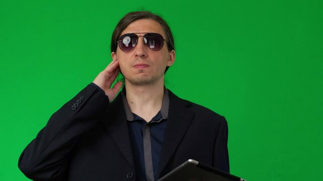 A mid-shot captures a serious and clandestine mood as a man in a suit and sunglasses, holding a tablet, touches his ear as if listening to an earpiece, against a green screen background