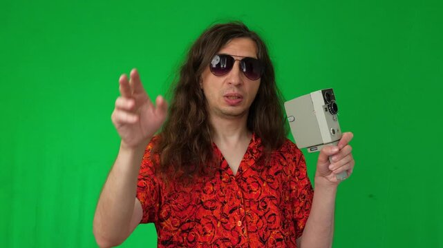 A mid-shot captures an active and creative mood as a man in sunglasses and a vibrant shirt holds a vintage film camera, extending his hand as if to frame a shot, against a green screen