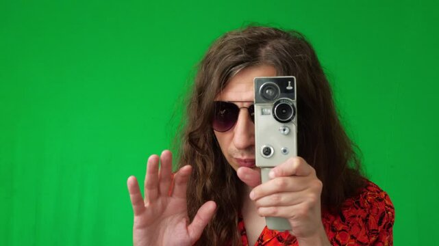 A mid-shot captures an active and creative mood as a man in sunglasses and a vibrant shirt holds a vintage film camera, extending his hand as if to frame a shot, against a green screen