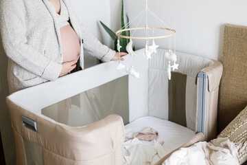 Pregnant woman holding baby bump and standing at baby crib with mobile, ultrasound picture, clothes and toy. Pregnancy. Baby room preparation and