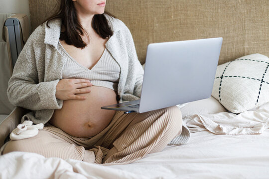 Pregnancy. Baby room preparation and online shopping. Pregnant woman holding laptop and hugging baby bump at baby crib with clothes and toy.