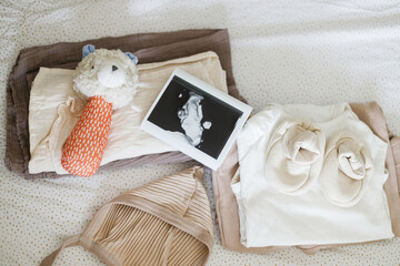 Pregnancy and getting ready for baby, top view. Modern baby crib with stylish baby clothes, toy, boho mobile, ultrasound picture.Preparation and making nursery