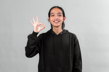 Young Asian person in black hoodie smiling and showing OK hand gesture, symbolizing agreement,...