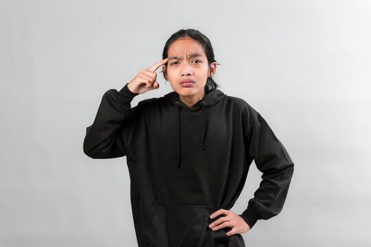 Young Asian man in black hoodie looking serious and confused, pointing at his head with index finger, expressing frustration or thinking hard, isolated on gray background.