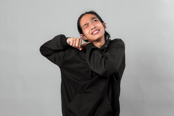 Asian man in a black hoodie grimacing with eyes closed, raising his fists and bending arms in discomfort, expressing pain or frustration against a neutral grey background.