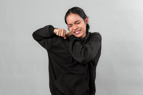Asian man in a black hoodie grimacing with eyes closed, raising his fists and bending arms in discomfort, expressing pain or frustration against a neutral grey background. - Powered by Adobe