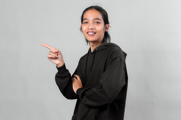 Asian man in black hoodie smiling confidently while pointing to the side with his index finger, standing against a plain gray background, promotional concept.