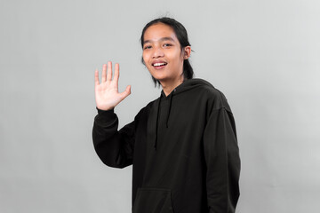 Young Asian man wearing a black hoodie smiling and waving hand while standing against a plain studio background. Friendly expression and welcoming gesture perfect for communication themes.